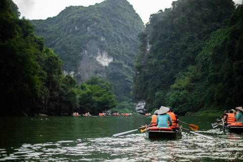       Rowers in conical hats paddle narrow boats through a lush limestone canyon on green water.
  