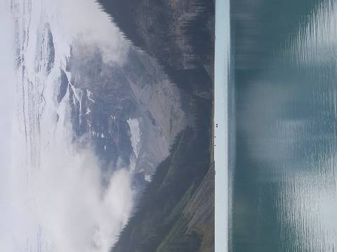       A turquoise glacial lake surrounded by steep forested slopes and mist-covered snowy peaks rising into the clouds.
  
