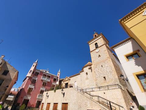       Upward view of a stone church tower with clock next to colorful historic buildings under a deep blue sky in Cieza.
  