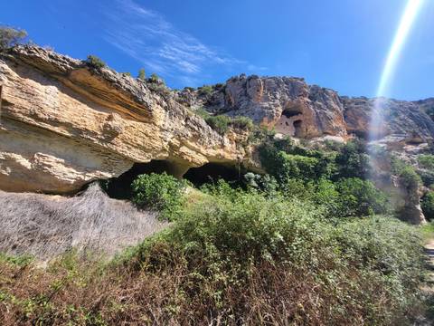       Rocky cliffside with natural caves and lush vegetation illuminated by a shaft of sunlight.
  