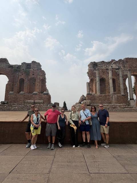       Group poses in front of the ancient brick arches of the Greek Theatre in Taormina under hazy skies.
  