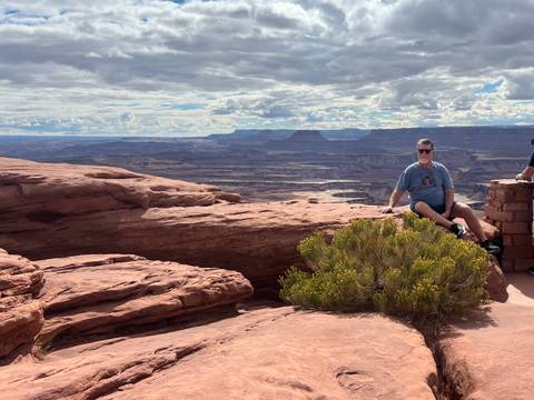       Traveler sitting on red rock ledge overlooking vast canyon landscape beneath partly cloudy sky
  