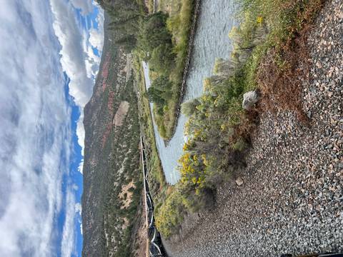       Curving river bordered by shrubs and wildflowers with distant hills under patchy clouds
  