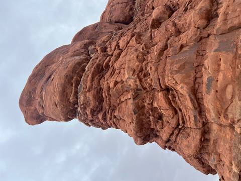       Tall, weather-sculpted red sandstone pillar reaching toward a grey sky in a desert landscape.
  