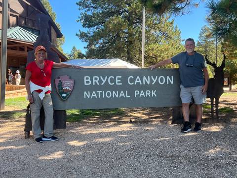       Two smiling travelers pose with the wooden entrance sign for Bryce Canyon National Park.
  