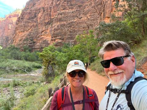       Smiling couple taking a selfie on a sandy canyon trail with towering walls behind.
  