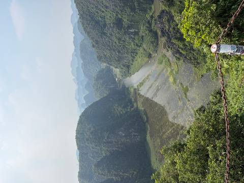       High viewpoint overlooking a lush river valley and layers of karst peaks fading into hazy distance.
  