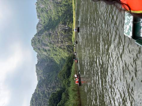       Long row of small paddle boats carrying tourists along a calm river framed by steep green cliffs.
  