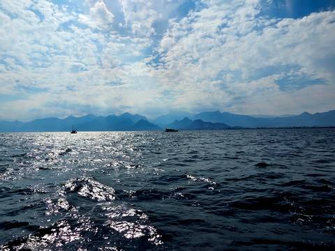       Sparkling Mediterranean sea with sailboats and hazy mountain range beyond under a cloud-dappled sky.
  
