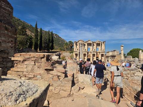       Tour group walks among the ruined walls and columns of ancient Ephesus under bright sky.
  