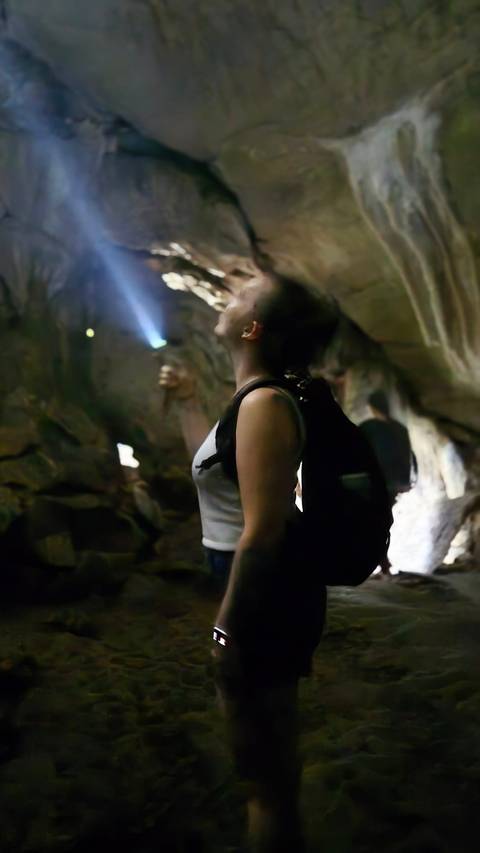       A backpacked traveller shines a flashlight upward while exploring a dim rocky cave passage.
  