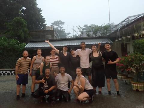       Smiling tour group poses outdoors in heavy rain, soaked but cheerful, raising arms in celebration.
  