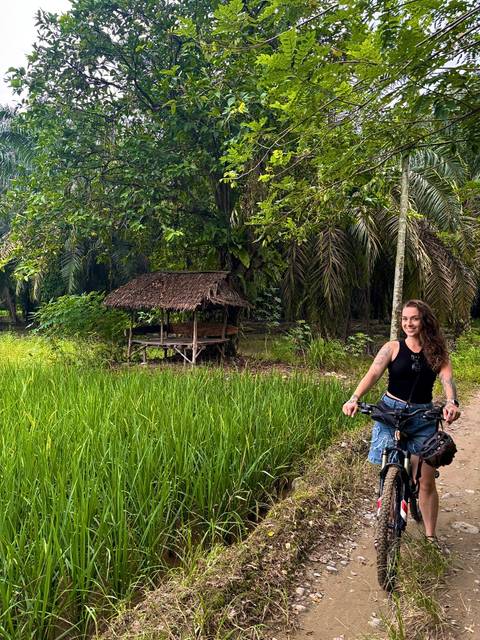       Woman on a bicycle rides past lush rice paddies and tall palms, smiling at the camera.
  