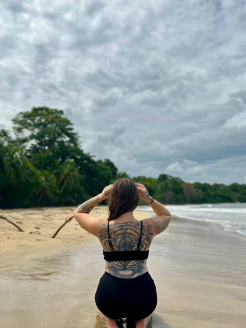       Tattooed woman gazes out at a secluded tropical beach bordered by lush jungle and cloudy skies.
  