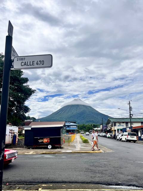       Arenal Volcano rises above the town of La Fortuna beneath scattered clouds, framed by a street sign and shops.
  