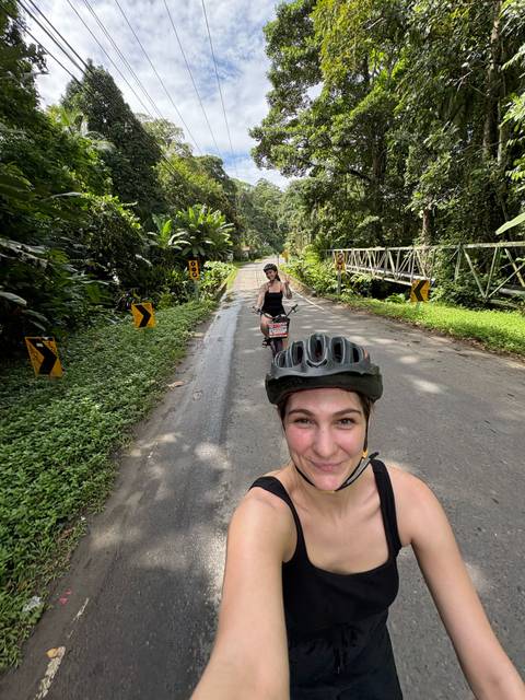       Selfie of two cyclists riding along a narrow jungle road with safety helmets and lush greenery all around.
  