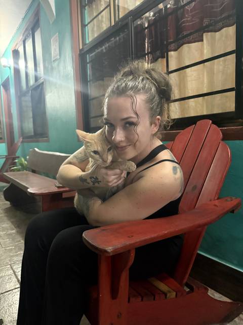       Traveler cuddles a ginger cat while seated on a red wooden chair in a cozy lodge.
  
