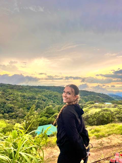       Smiling traveler with braided hair stands before a golden sunset over verdant hills.
  