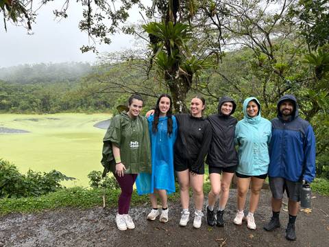       Rain-soaked group in ponchos smiles in front of a vivid green crater lake and misty hills.
  