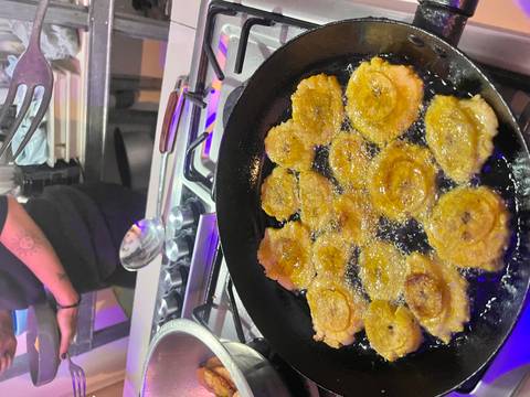       Slices of fried plantain sizzle in oil inside a cast-iron skillet on a stovetop.
  