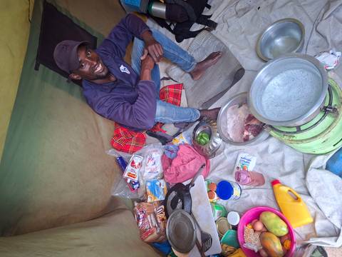       Camp cook smiling inside a tent surrounded by food supplies and cooking utensils laid out on fabric.
  