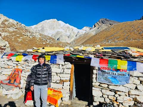       Local tea-house owner poses outside a stone hut draped with prayer flags against snowy peaks.
  