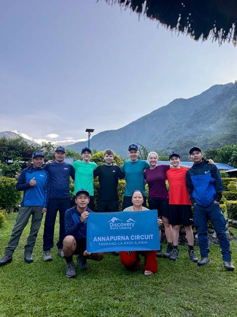       A trekking team poses with a banner in a lush garden with Himalayan peaks just visible through cloud.
  