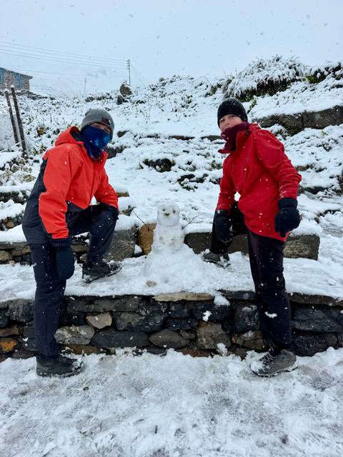       Two trekkers kneel beside a small snowman during a gentle snowfall on a stone terrace.
  