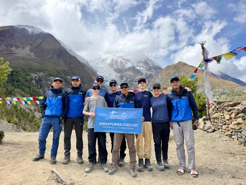       A trekking group celebrates at a high-altitude pass holding an Annapurna Circuit banner with rugged peaks behind.
  