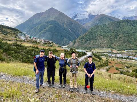       A family of trekkers smiles on a grassy ridge overlooking a broad Himalayan valley dotted with villages.
  