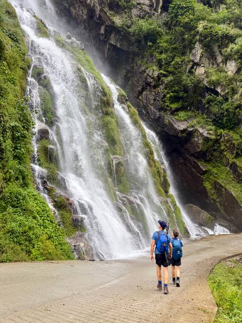       A tall, multi-tiered waterfall plunges down a mossy cliff while a lone hiker gazes up from the base.
  