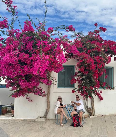       Bright pink bougainvillea drapes over a whitewashed Greek-style house while two travelers sit below the blossoms
  