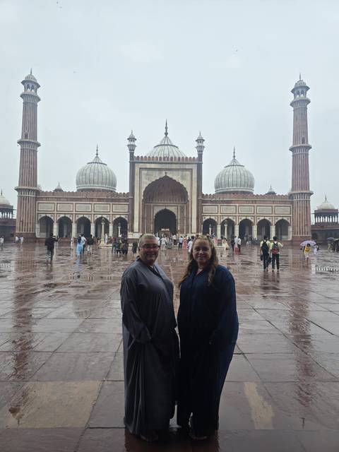       Two visitors in modest attire stand on the wet marble courtyard of Jama Masjid with its domes and minarets rising behind.
  