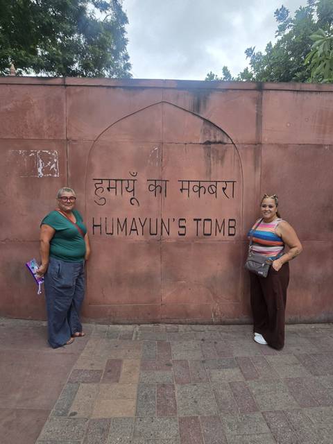       Travellers smile beside a sandstone wall inscribed with “Humayun’s Tomb” in Hindi and English.
  