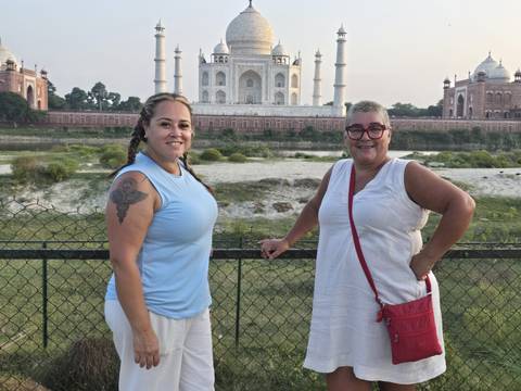       Two women pose across the river with the Taj Mahal glowing softly in the background at dusk.
  