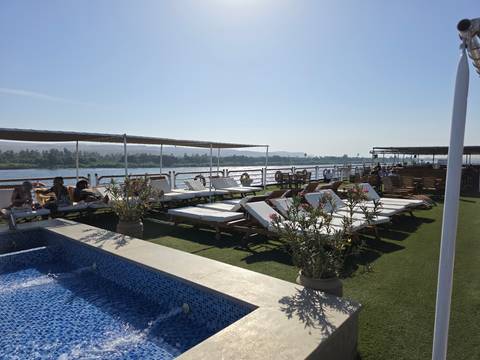       Sun-drenched sundeck of a Nile cruise ship with loungers, small pool and river scenery drifting past.
  