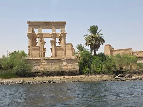       Ancient sandstone columns and architraves of Philae Temple rise above lush riverside vegetation.
  
