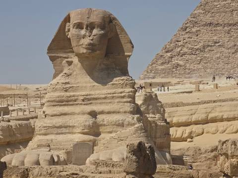       Close-up of the Great Sphinx’s weathered face with a pyramid and desert plateau behind.
  