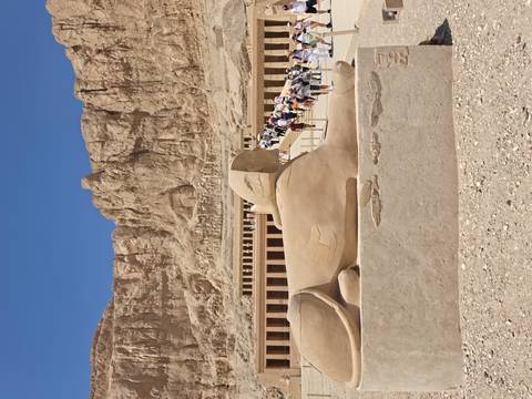       Stone sphinx statue in front of the terraced colonnades of Hatshepsut’s Temple set against rugged cliffs.
  