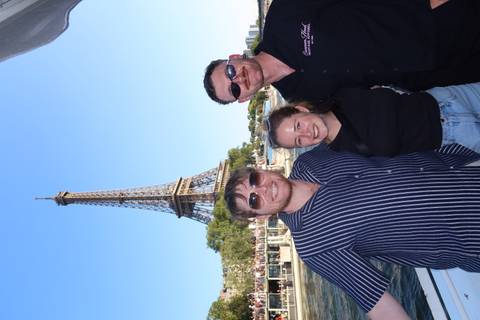       Three friends smile on a boat cruise with the Eiffel Tower towering behind them on a sunny afternoon.
  