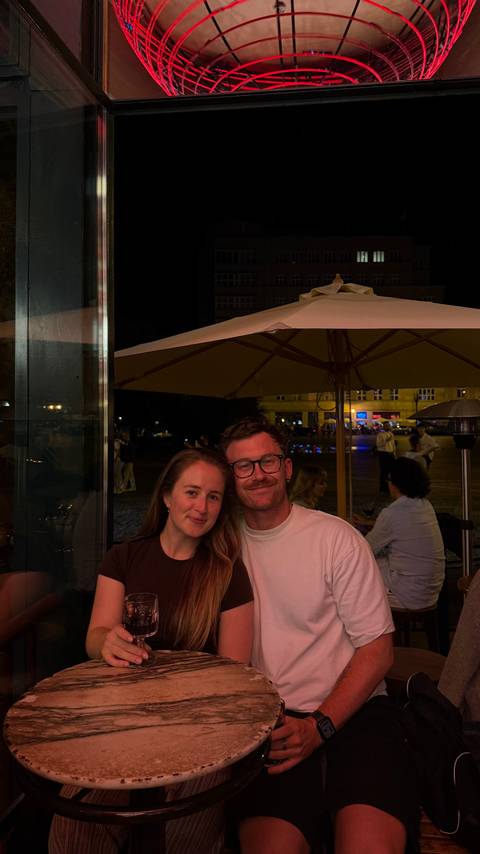       Couple share a table under a street-side umbrella at night with blurred city lights behind.
  