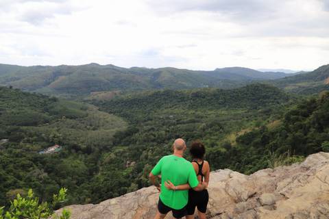       Couple sits on rocky outcrop admiring a vast green valley and distant mountains.
  