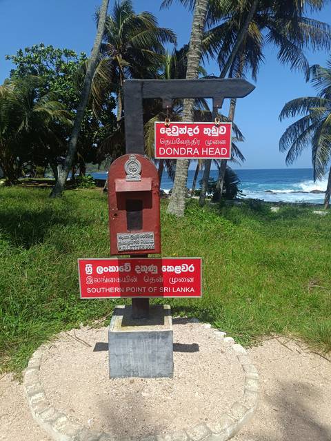       Red signposts mark Dondra Head, the southernmost point of Sri Lanka, with palm trees and turquoise sea beyond.
  