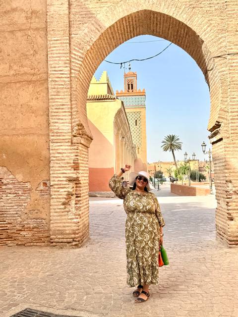      Woman in patterned dress poses beside sunlit adobe walls and palm in Marrakesh.
  