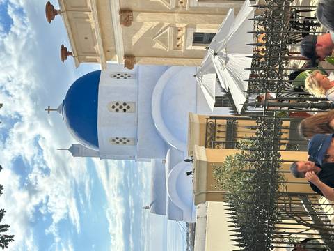       Blue-domed white church of Santorini rises above a busy terrace of café umbrellas and tourists.
  