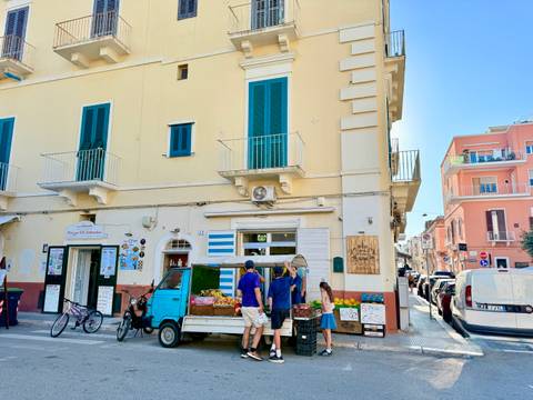       Locals buy produce from a small blue three-wheeled truck outside pastel buildings.
  