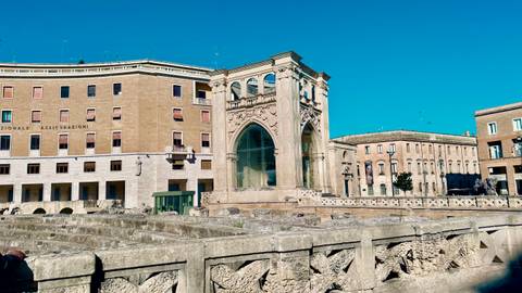       Ancient Roman amphitheater ruins surrounded by 20th-century buildings in a sunny square.
  