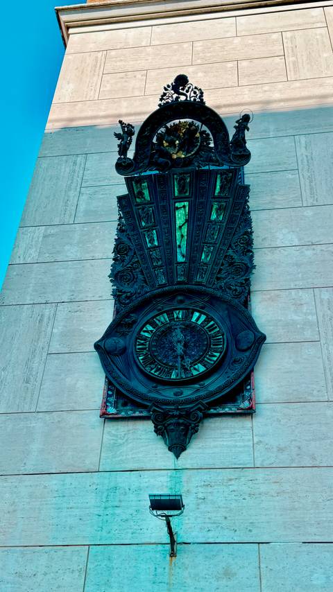       Close-up of an ornate bronze clock with Roman numerals mounted on a stone wall.
  