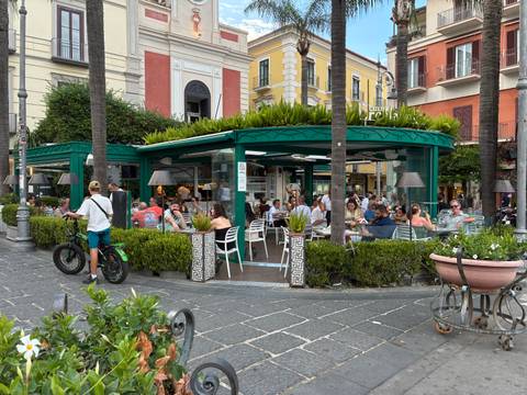       Open-air café in a lively Italian piazza with many patrons socialising.
  