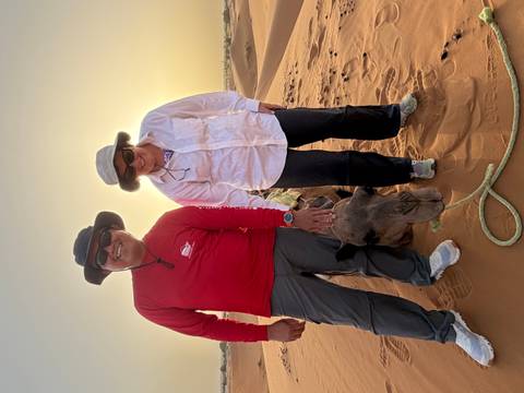       Smiling travellers with a camel at sunset on desert dunes.
  